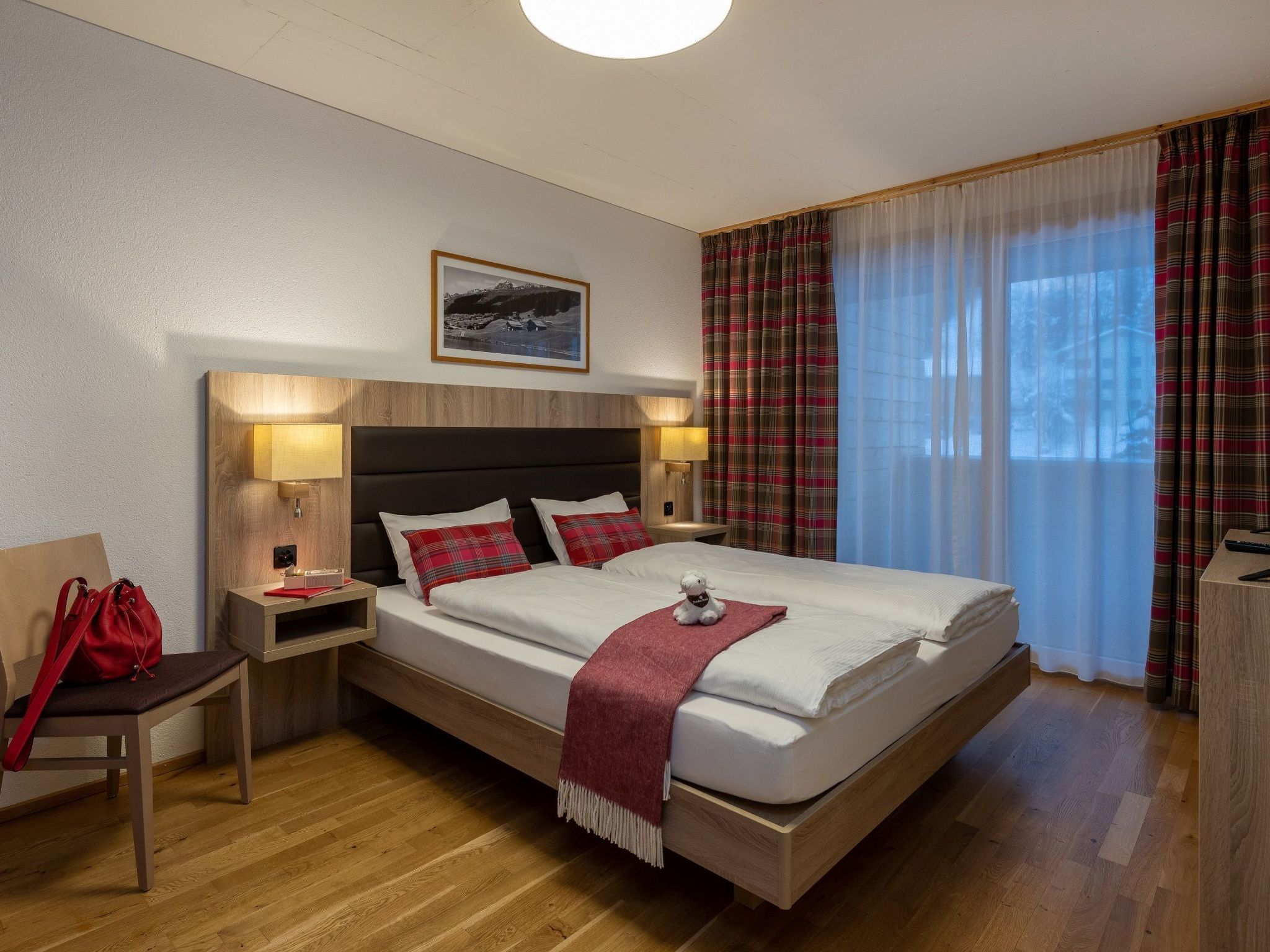 Ein Hotelzimmer mit einem Bett, Nachttischlampen, einer roten Tasche, einem Sessel, einem Schreibtisch und einem Fenster mit Vorhängen.