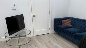 Una habitación con una televisión de pantalla plana sobre una mesa de cristal, una puerta blanca cerrada, un sofá de terciopelo azul con una manta marrón y suelo de madera clara.