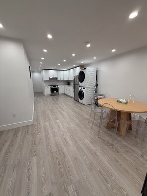 A modern kitchen with white cabinets, a built-in oven, a washing machine, and a dining area with a wooden table and clear chairs.