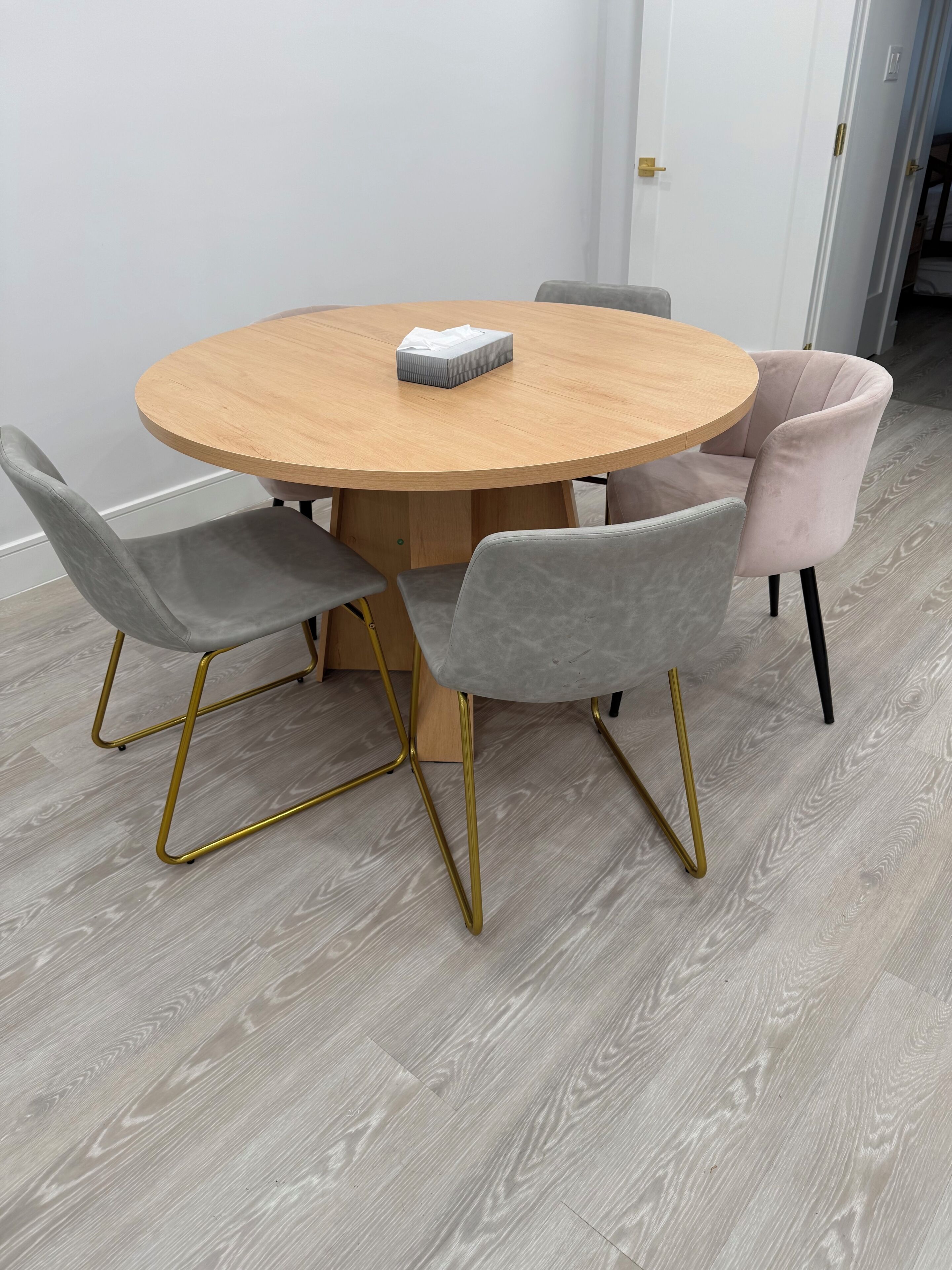 Une table ronde en bois, accompagnée de quatre chaises : l’une avec un cadre doré et les trois autres recouvertes d’un tissu gris.