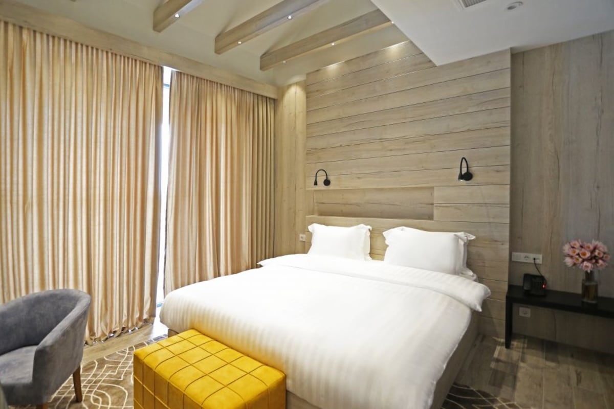 Una habitación de hotel moderna con una cama grande, un cabecero de madera, una mesita de noche con una lámpara, una silla y una pequeña mesa con un jarrón de flores.