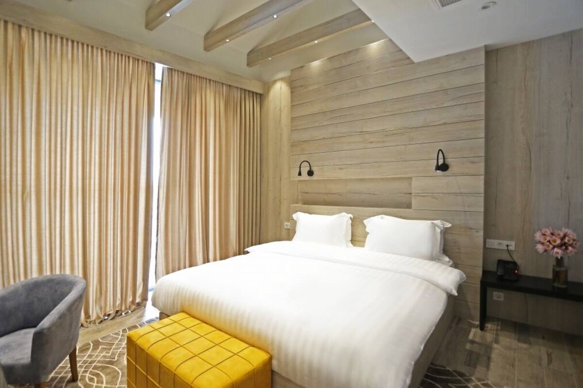 Una habitación de hotel moderna con una cama grande, un cabecero de madera, una mesita de noche con una lámpara, una silla y una pequeña mesa con un jarrón de flores.