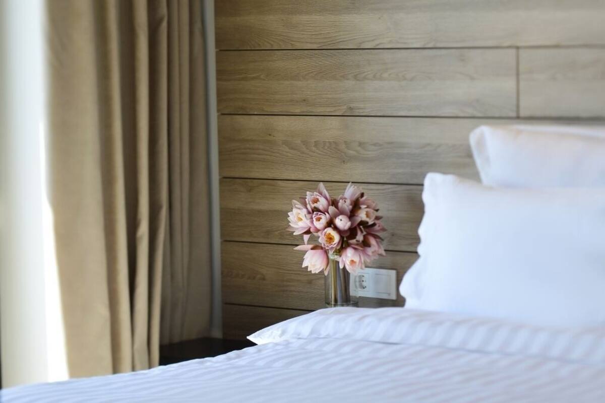 Habitación de hotel con cabecera de madera, cama blanca con almohadas y un florero con flores rosas sobre una mesita de noche.