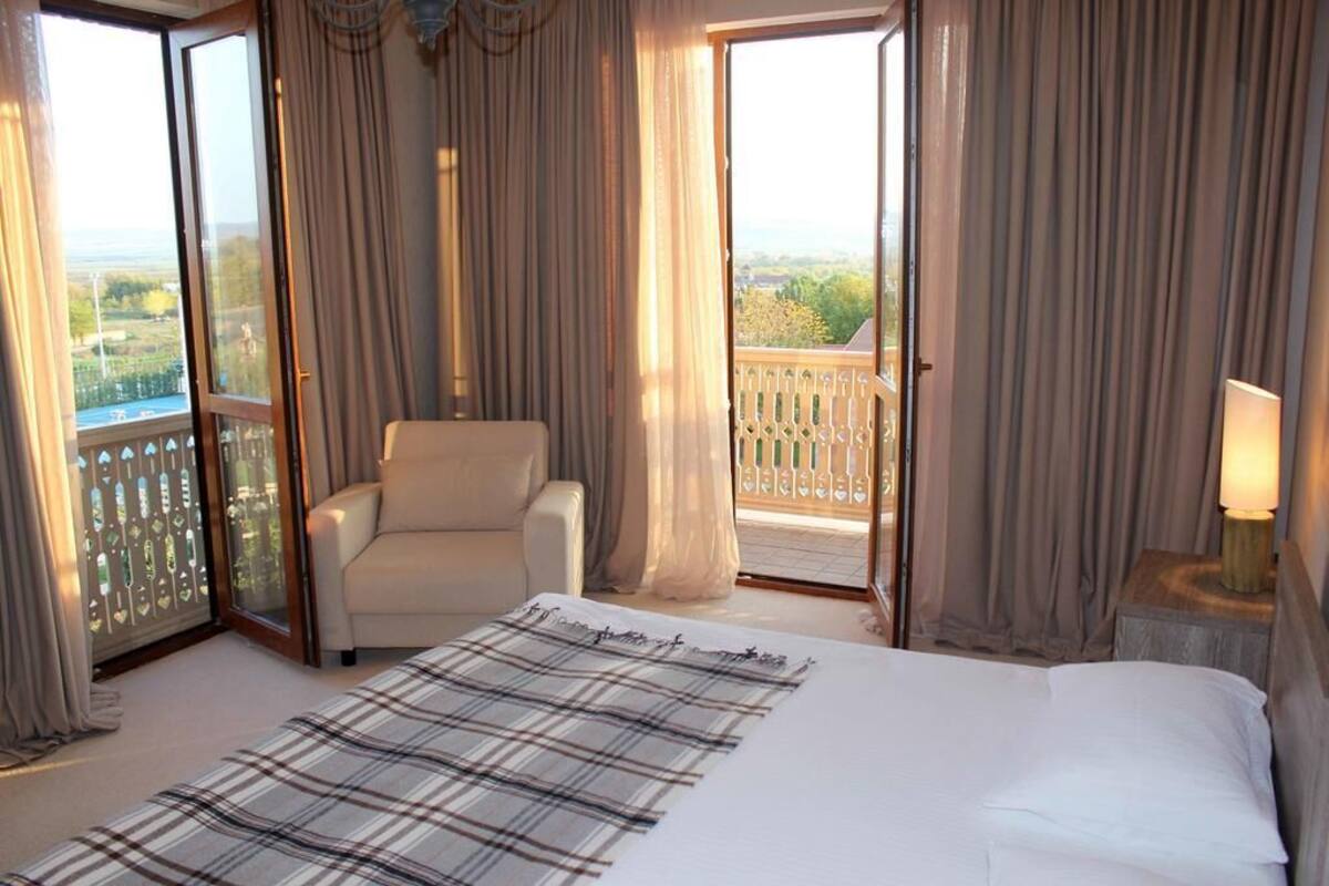Habitación de hotel con cama, una silla, mesita de noche con lámpara y balcón con cortinas.