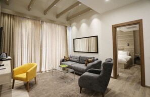 A modern living room with a sofa, armchairs, a coffee table, and a TV. There is a bed visible through an open door. - Ambassadori Kachreti Golf Resort (Gurjaani)