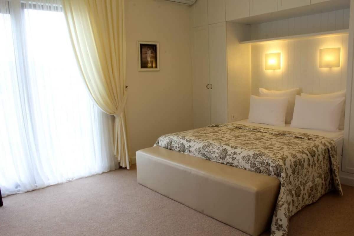 A bedroom with a bed, bench, curtains, wall-mounted lights, and a framed picture.