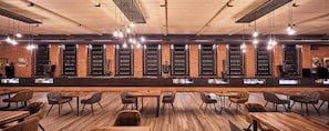 Una zona de bar con mesas y sillas de madera, una estantería con botellas de vino y lámparas colgantes.