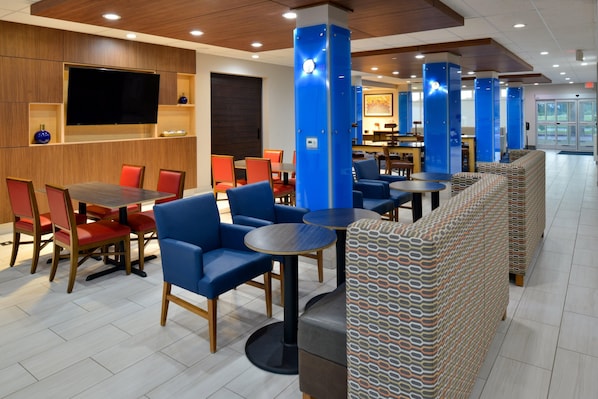 A modern hotel lobby with a large flat-screen TV, multiple seating areas with blue and patterned furniture, and a tiled floor.