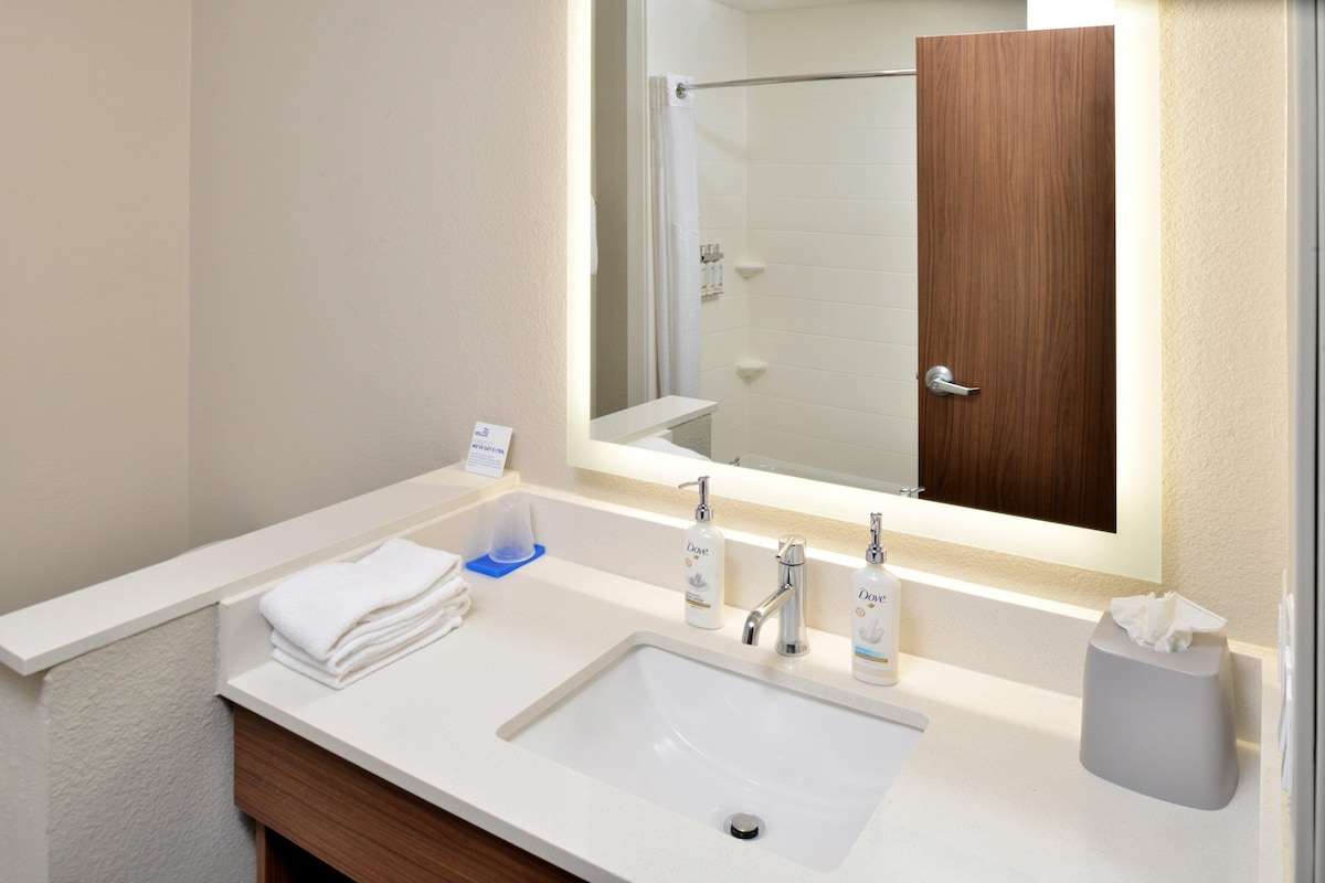 Une salle de bain d’hôtel avec un lavabo blanc, deux robinets, un miroir et une porte en bois.