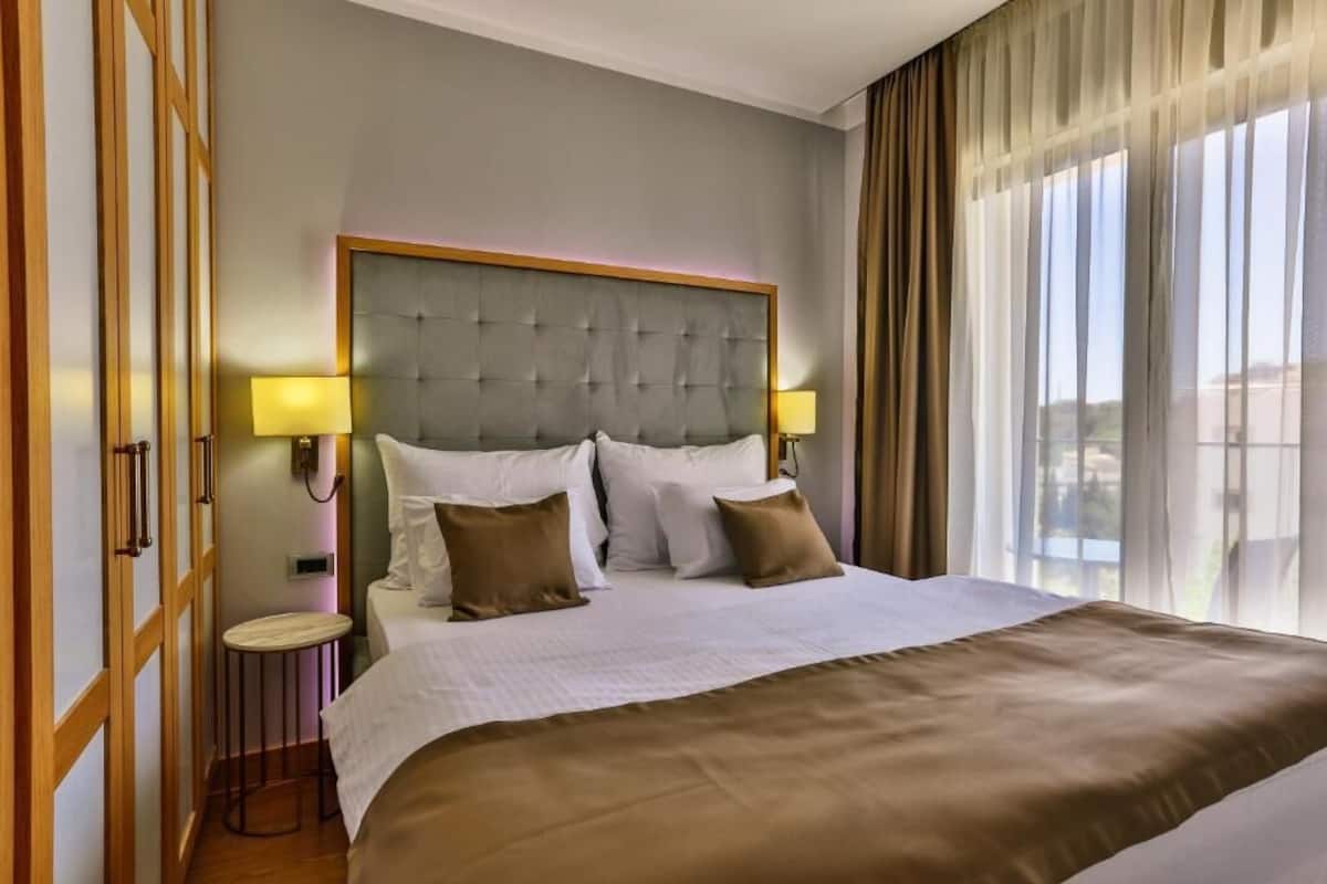 Une chambre d’hôtel avec un grand lit, deux lampes de chevet, une table de chevet en bois et une vue sur l’extérieur à travers des rideaux légers.