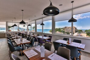 A restaurant with multiple tables and chairs, large windows offering a view of the sea and buildings.