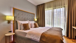 A neatly made bed with a grey headboard, two bedside lamps, and a view of the outdoors through sheer curtains.