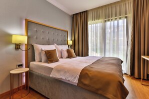 A neatly made bed with a grey headboard, two bedside lamps, and a view of the outdoors through sheer curtains.