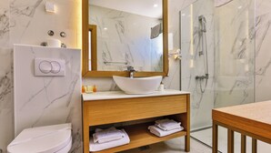 A modern bathroom with a marble wall, a wooden vanity, a white sink, a toilet, and a glass shower enclosure.