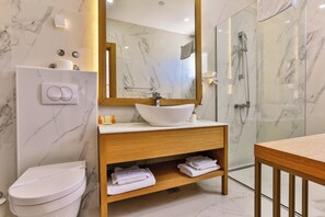 A modern bathroom with a marble wall, a wooden vanity, a white sink, a toilet, and a glass shower enclosure.