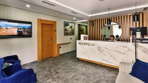 A hotel reception area with a marble reception desk, a television mounted on the wall, and a seating area with blue chairs.
