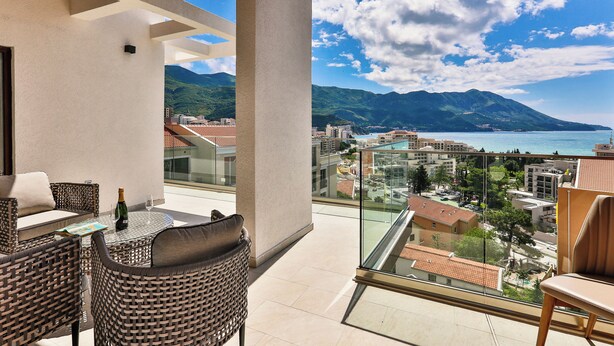 A balcony with wicker furniture, a bottle of champagne, and a glass. It overlooks a coastal city with mountains in the distance.