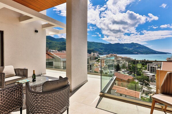 A balcony with wicker furniture, a bottle of champagne, and a glass. It overlooks a coastal city with mountains in the distance.