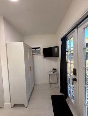 Ein kompakter Raum mit einem weißen Kleiderschrank, einem kleinen Tisch mit Pflanze, einem an der Wand befestigten Fernseher und einer Glastür zum Außenbereich.