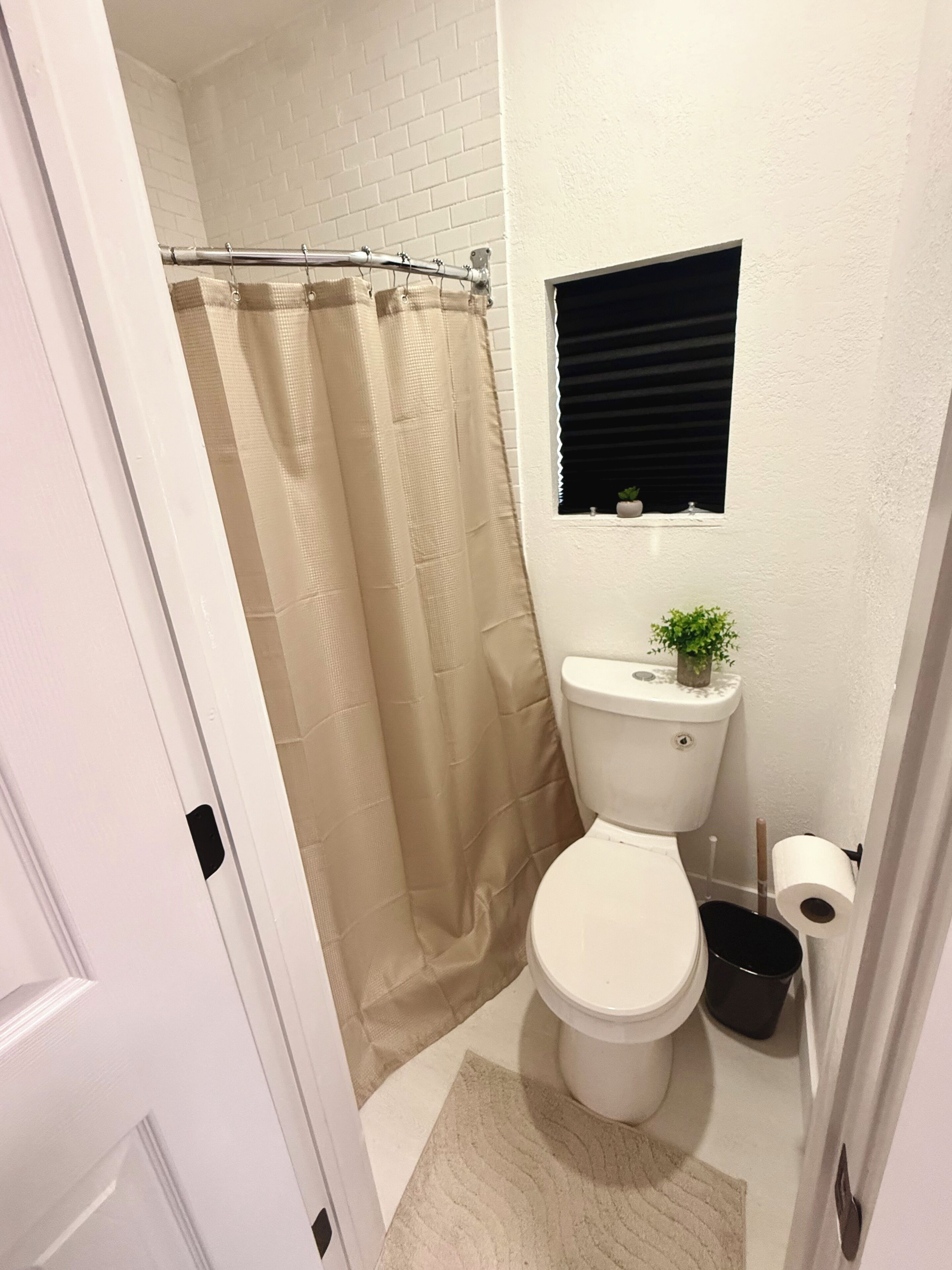 Une salle de bain équipée de toilettes, d’un rideau de douche, d’une fenêtre avec des stores, d’une petite plante en pot et d’un porte-rouleau de papier toilette.