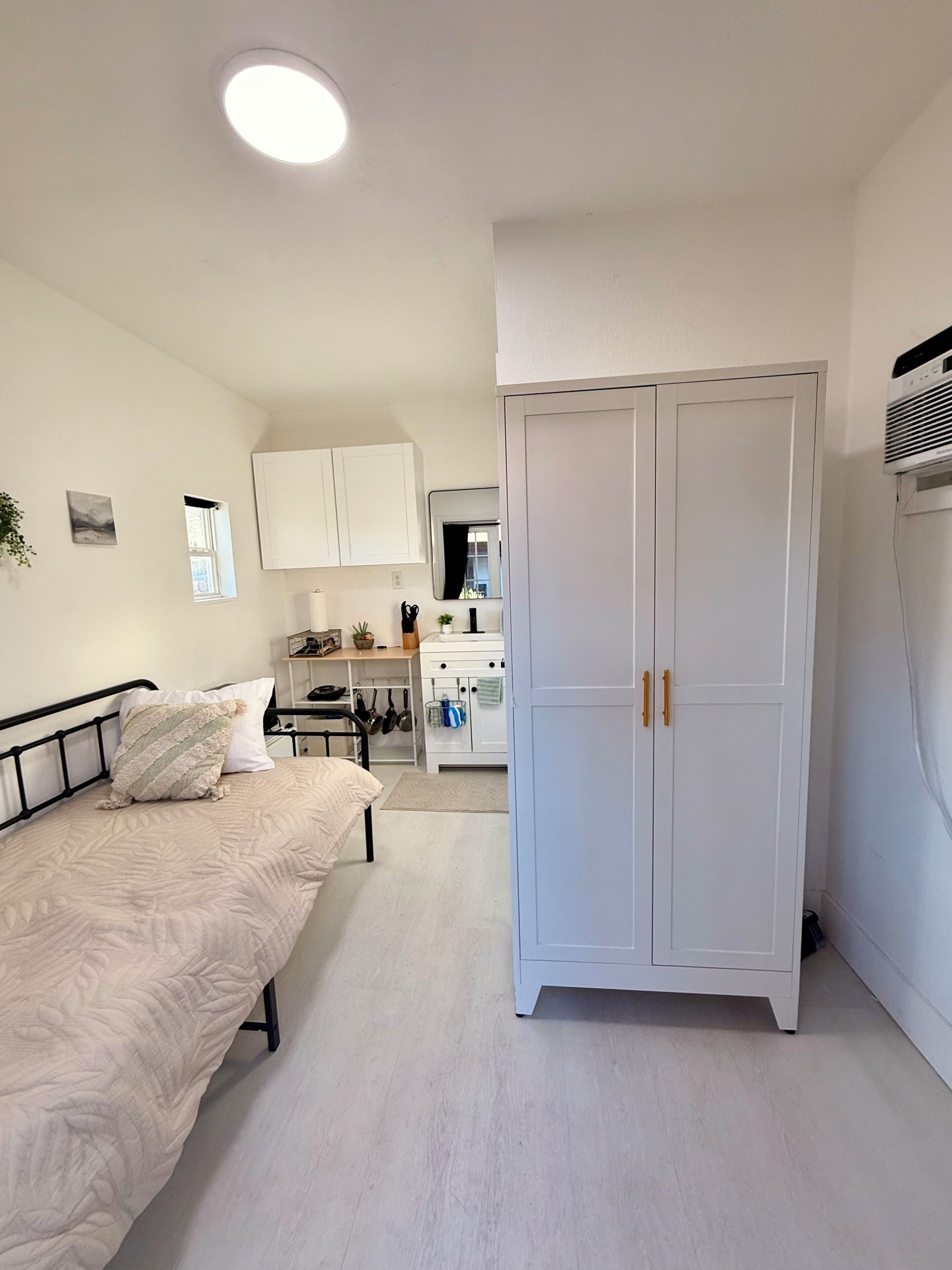 A bedroom with a bed, a wardrobe, and a kitchenette in the background.