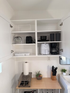 Eine kleine Küche mit weißen Schränken, einer Kaffeemaschine, einem Toaster und einer Arbeitsplatte mit Pflanze und Besteck.