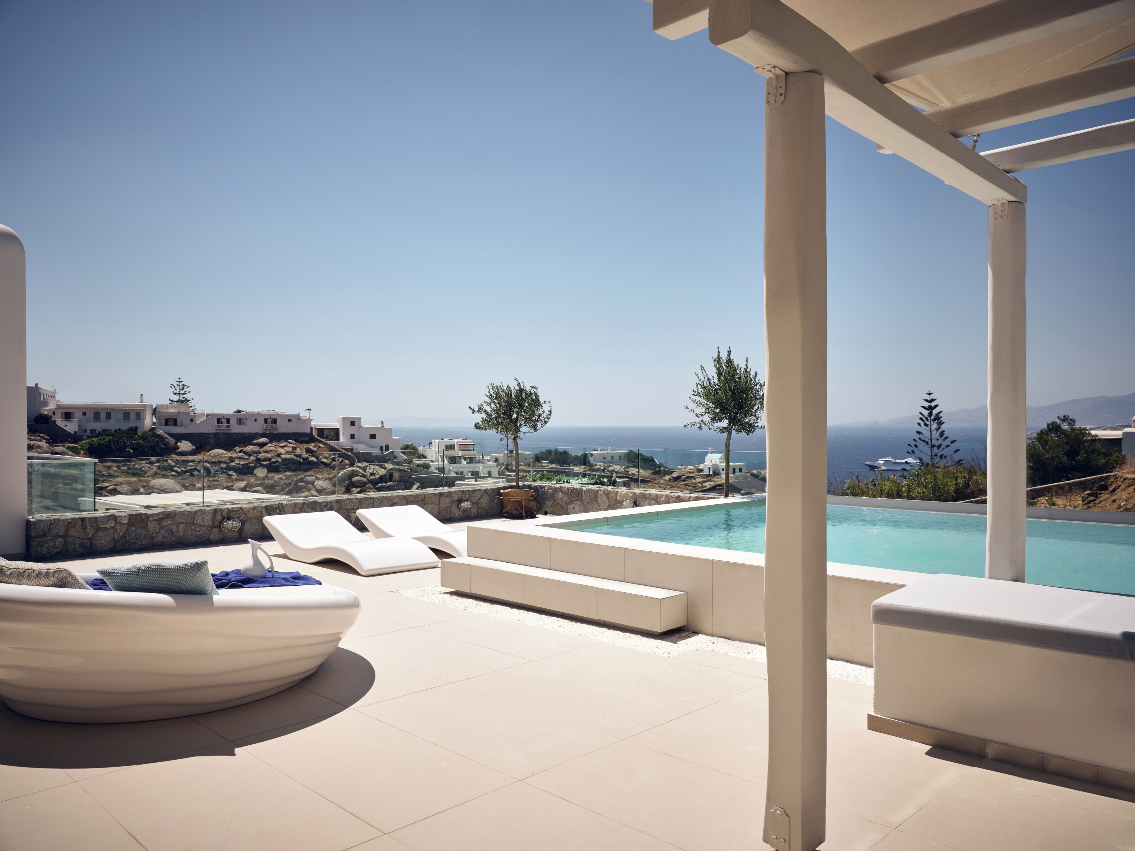 A poolside area with lounge chairs, a stone wall, and a view of the sea and buildings.