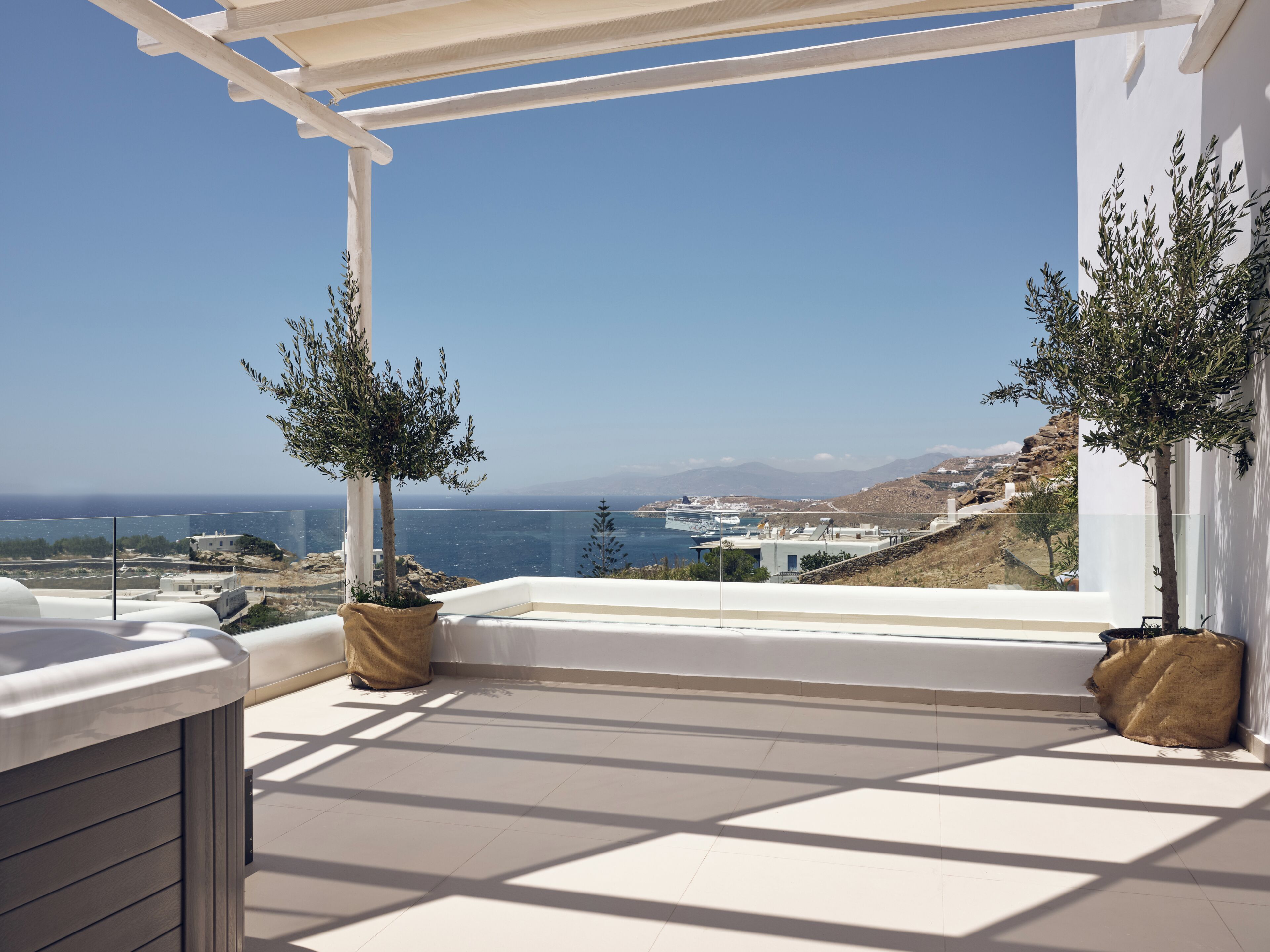 A balcony with a view of the sea and a cruise ship, potted olive trees, and a white pergola.