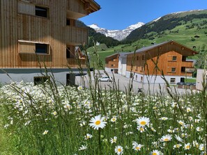 Un bâtiment moderne en bois, à plusieurs étages, avec un espace de stationnement et, au premier plan, un champ de marguerites.