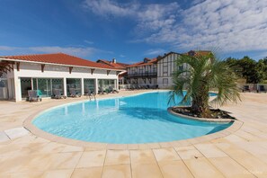 A swimming pool with lounge chairs, a palm tree, and buildings in the background. - Olydea la Chalosse (Cassen)