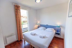 A bedroom with a bed, two bedside tables with lamps, a window with curtains, and a radiator. - Olydea la Chalosse (Cassen)