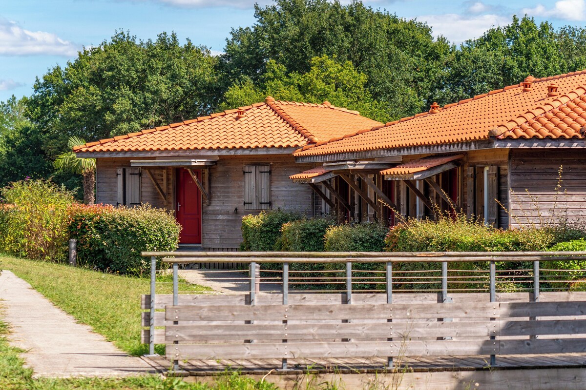 A rustic wooden building with a red door and a tiled roof, surrounded by greenery and a wooden fence.
