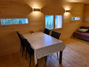 Een houten hut met een eettafel, stoelen en twee ramen met uitzicht op bomen en een besneeerd landschap.