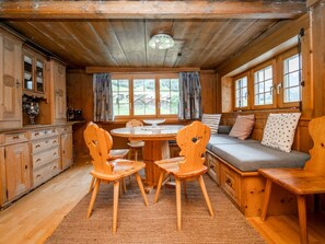 Interior acolhedor de cabana com mesa de jantar de madeira, duas cadeiras, um banco e um sofá com almofadas.