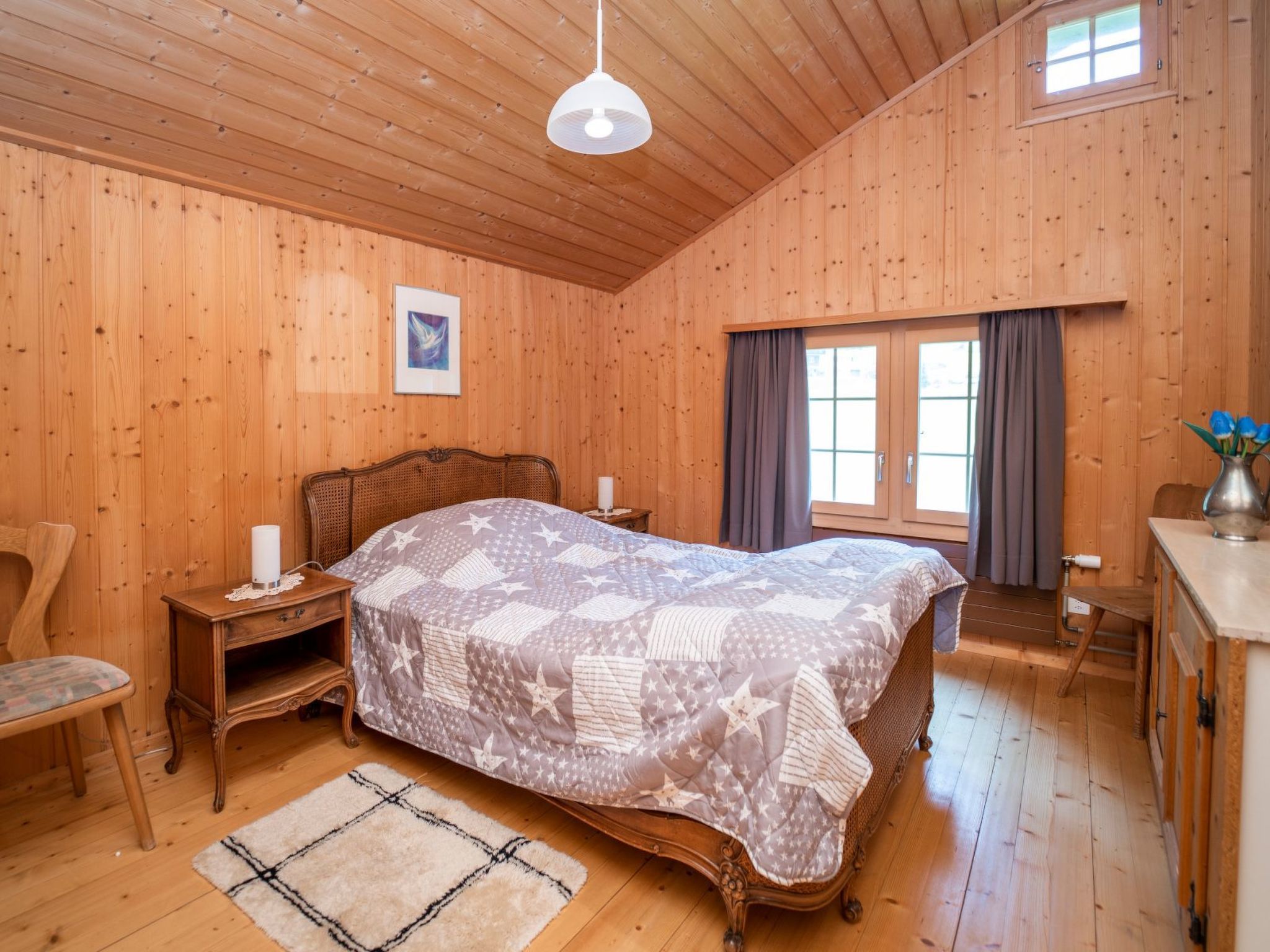 A wooden-paneled bedroom with a bed, a chair, a nightstand, a window with curtains, and a small table with a vase.