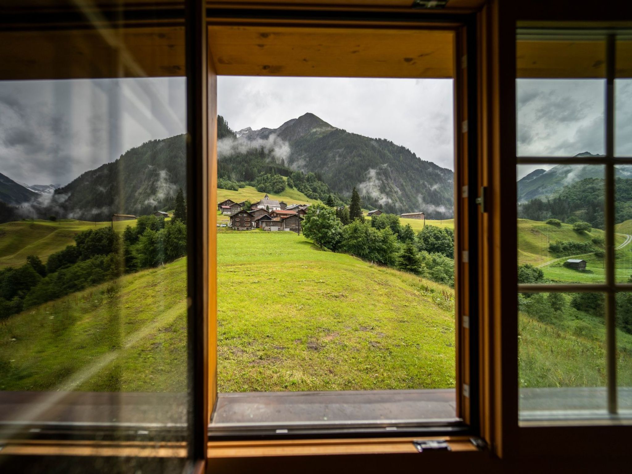 Vista a través de una ventana de un paisaje montañoso con una casa en primer plano.