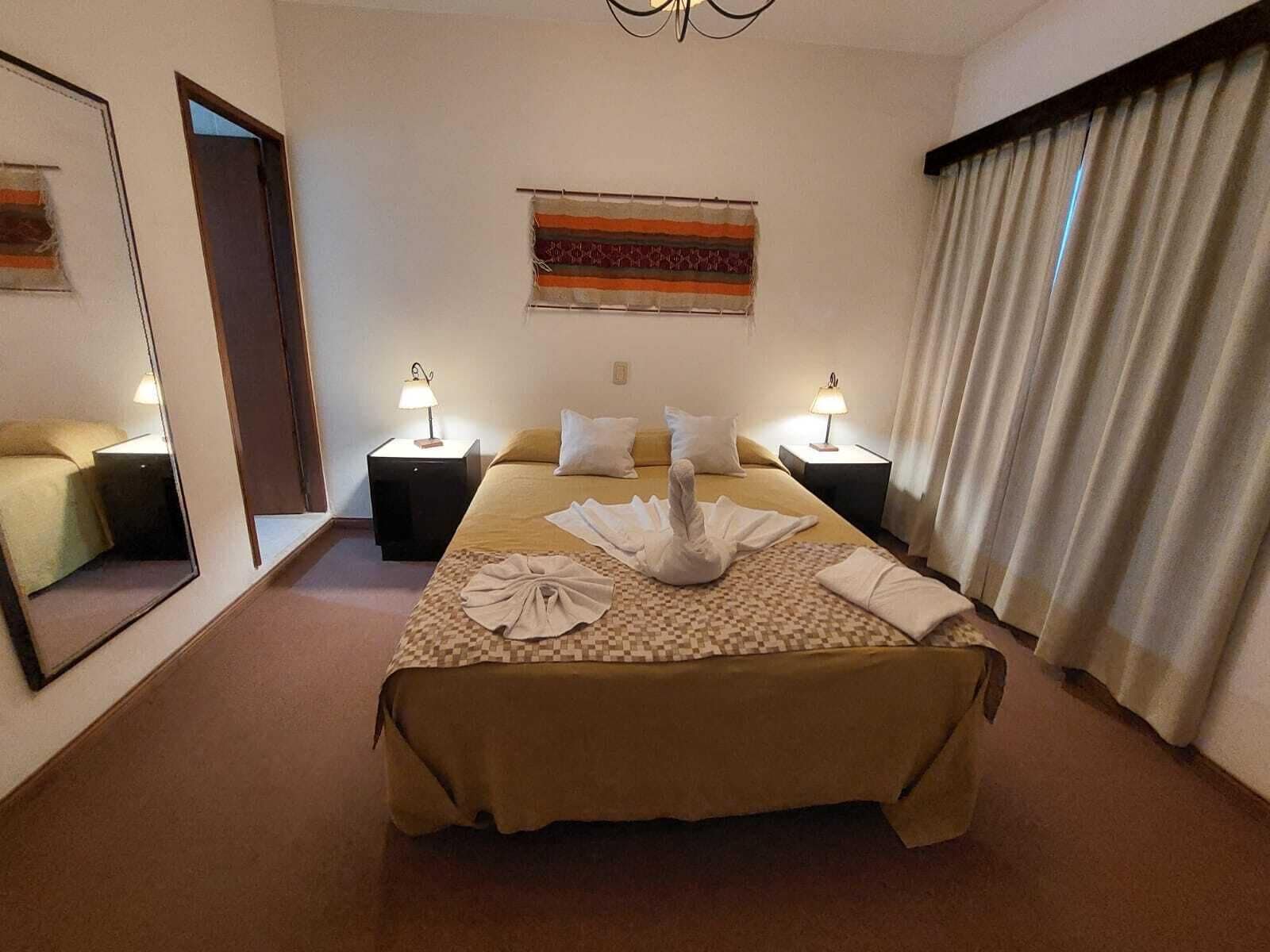 Een hotelkamer met een groot bed, twee nachtkastjes met lampen, een spiegel en een raam met gordijnen.