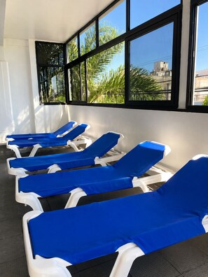 Una fila de tumbonas azules con estructura blanca, dispuestas en una sala de estar con grandes ventanales.