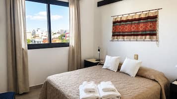 A hotel room with a bed, two pillows, a window with curtains, a wall-mounted tapestry, and a bedside table with a lamp.