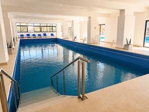 An indoor swimming pool with a clear view of the pool area, a set of stairs leading into the pool, and a seating area with chairs.