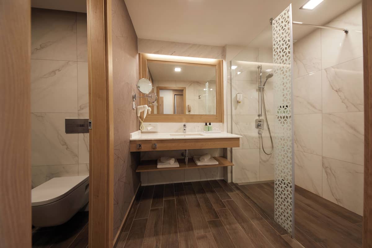 Une salle de bain moderne avec un meuble-lavabo en bois, un grand miroir, des toilettes et une douche à l’italienne agrémentée d’un panneau décoratif.
