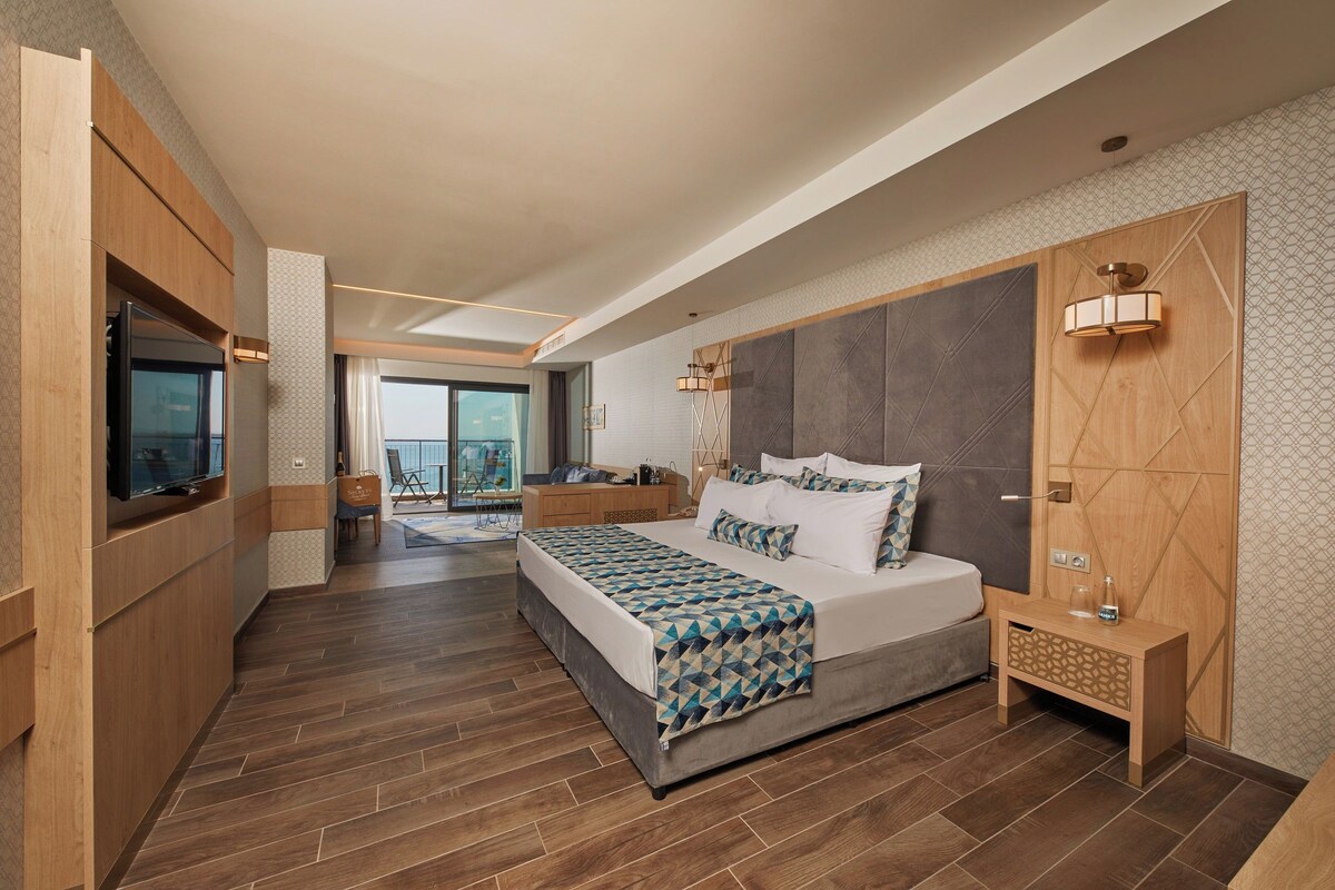 A modern hotel room with a large bed, a TV, a wooden nightstand, and a view of the city.