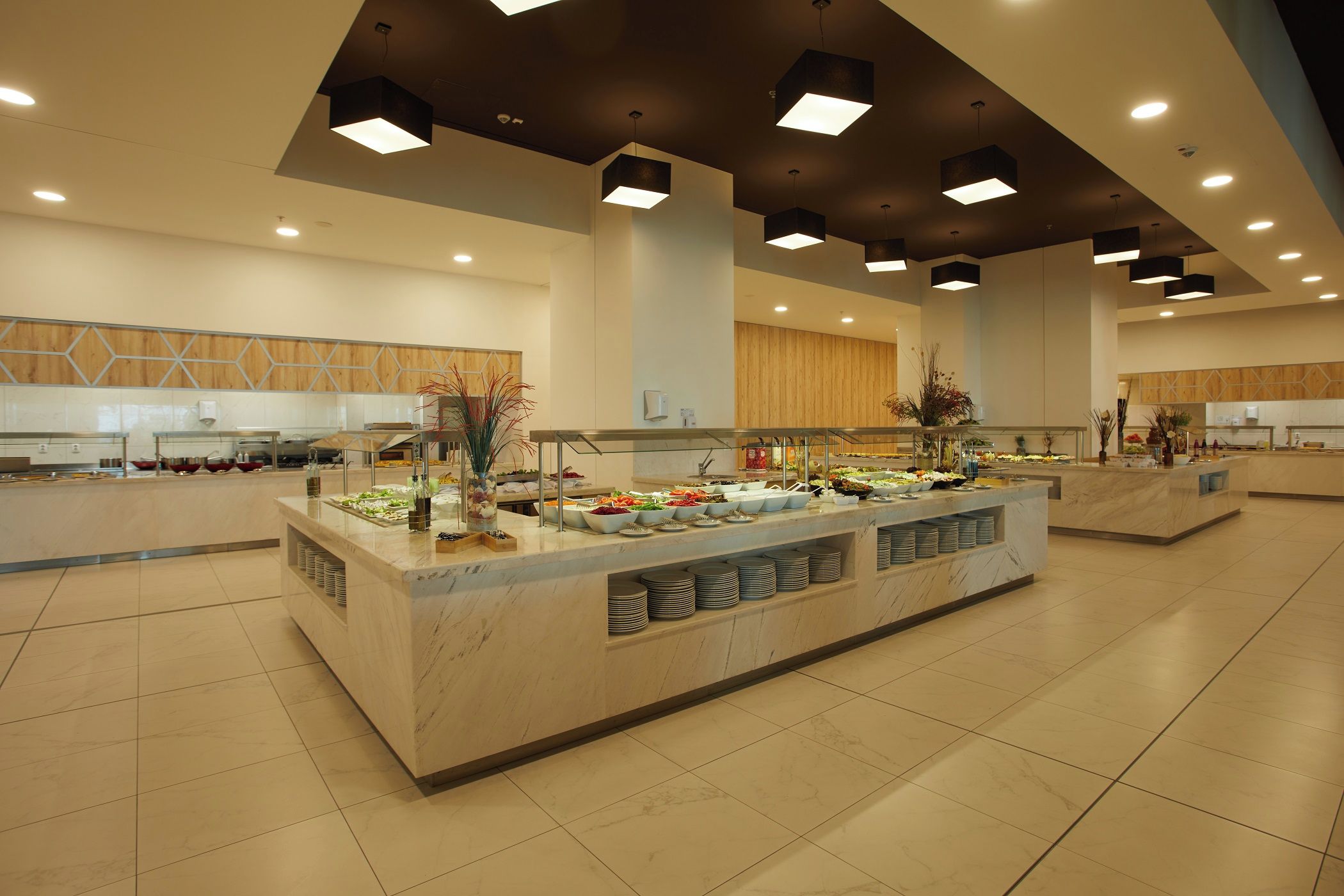 Amplia zona de buffet con múltiples estaciones de servicio, suelo de baldosas e iluminación colgante moderna.