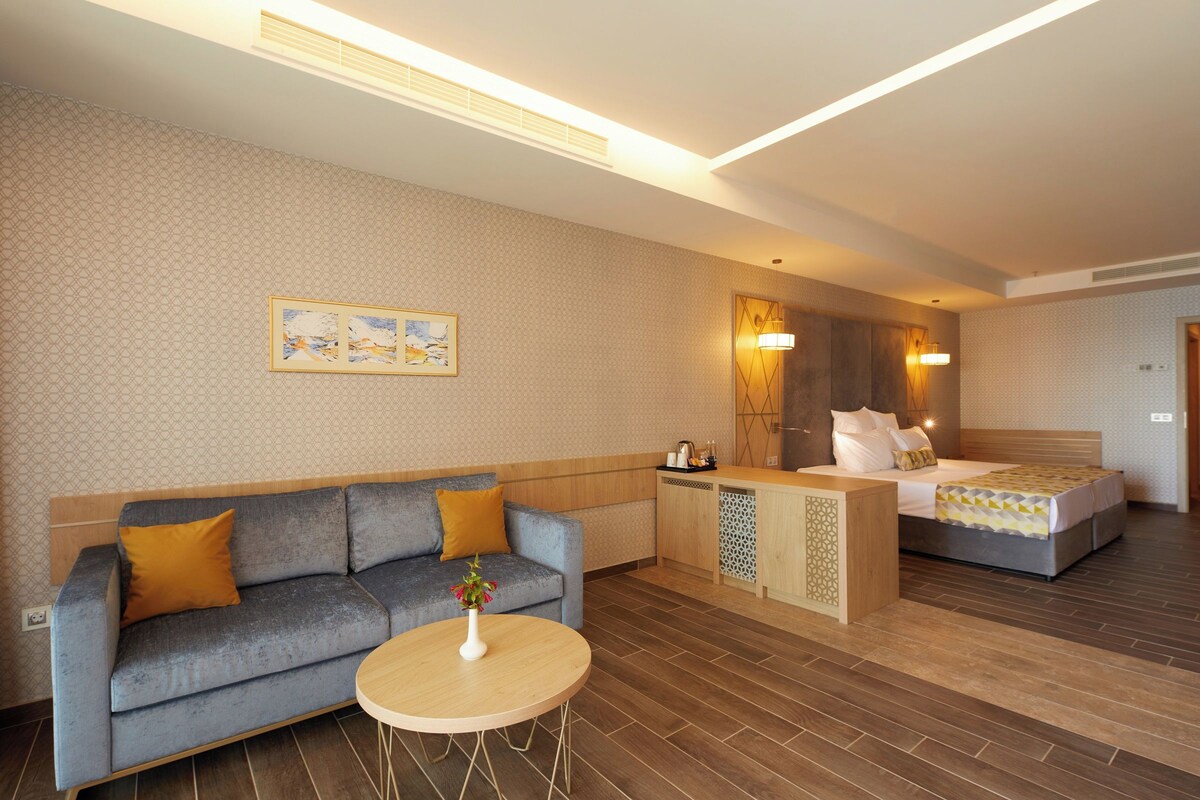 A modern hotel room with a sofa, a small round table, a bed, and a kitchenette area.
