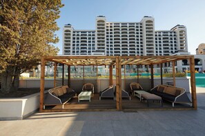 A covered outdoor seating area with modern furniture, a tree, and a large building in the background. - Secrets Sunny Beach Resort & Spa (Nessebar)