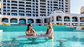 Piscina com duas pessoas sentadas numa mesa, um grande edifício de hotel ao fundo.