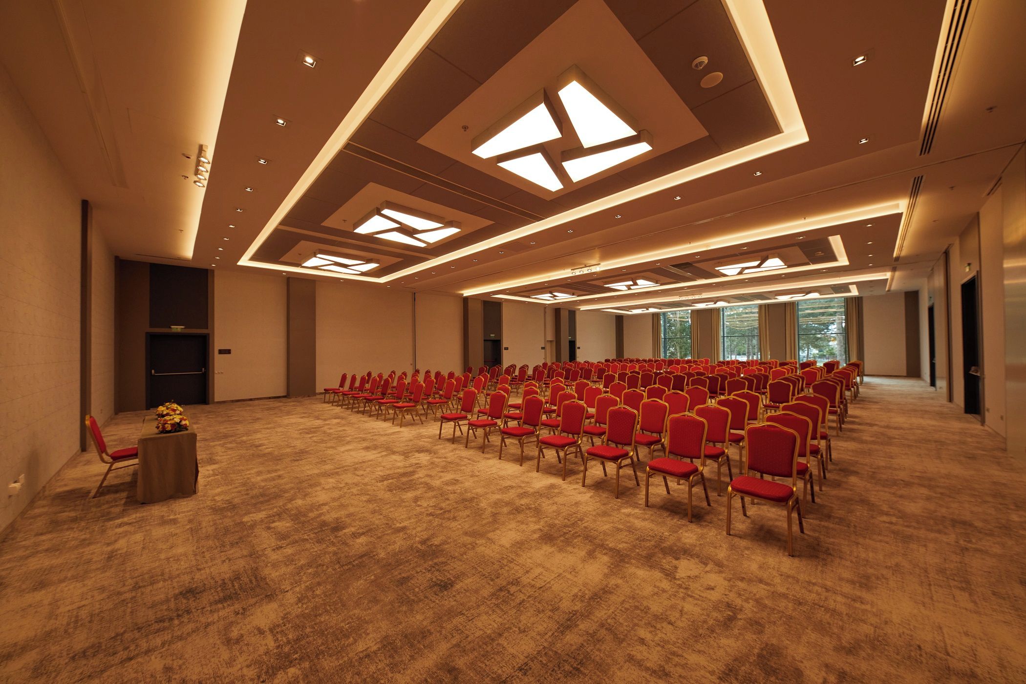Un salón de conferencias con hileras de sillas rojas, piso alfombrado e iluminación empotrada en el techo.