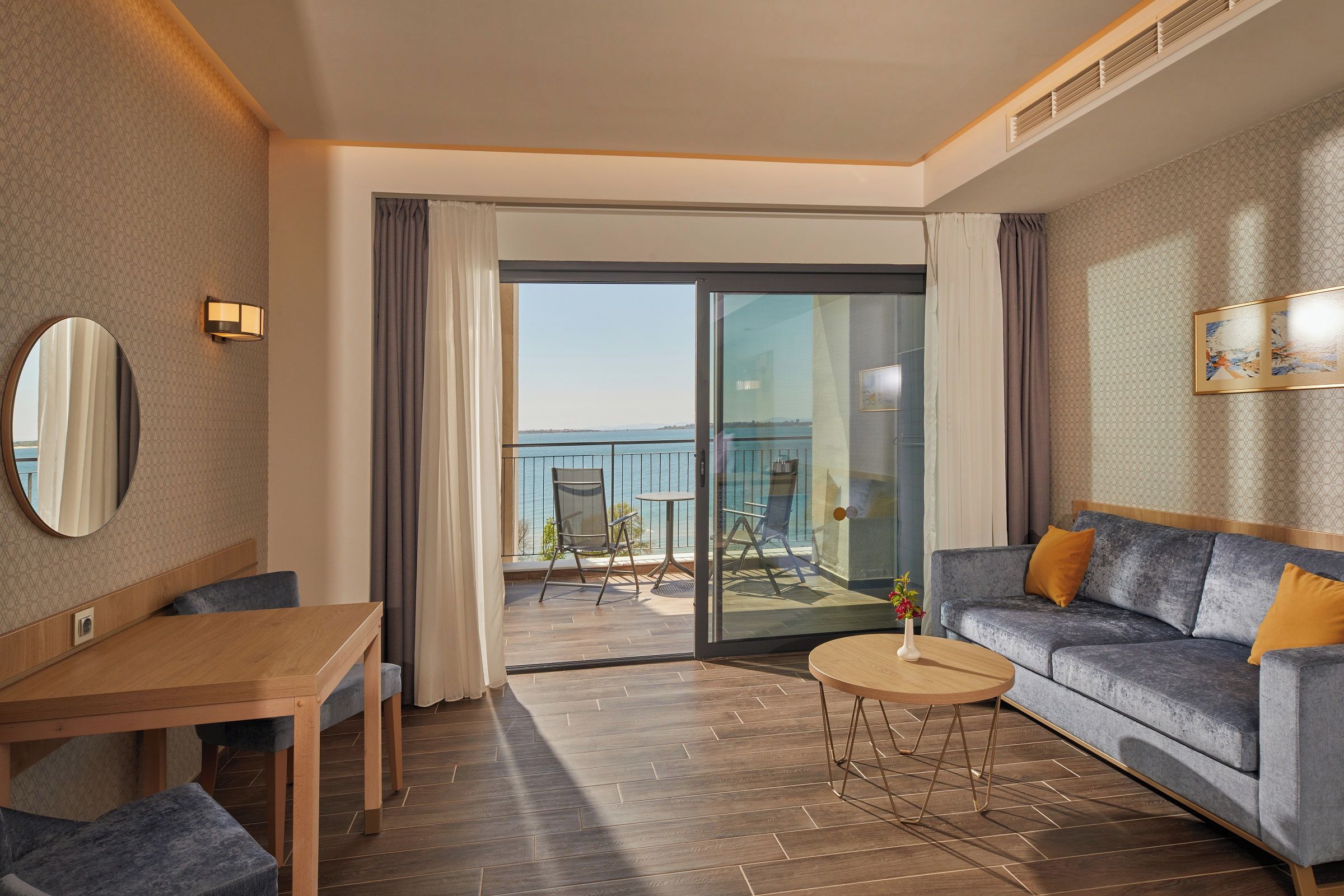 A hotel room with a balcony, a sofa, a dining table, and a mirror.