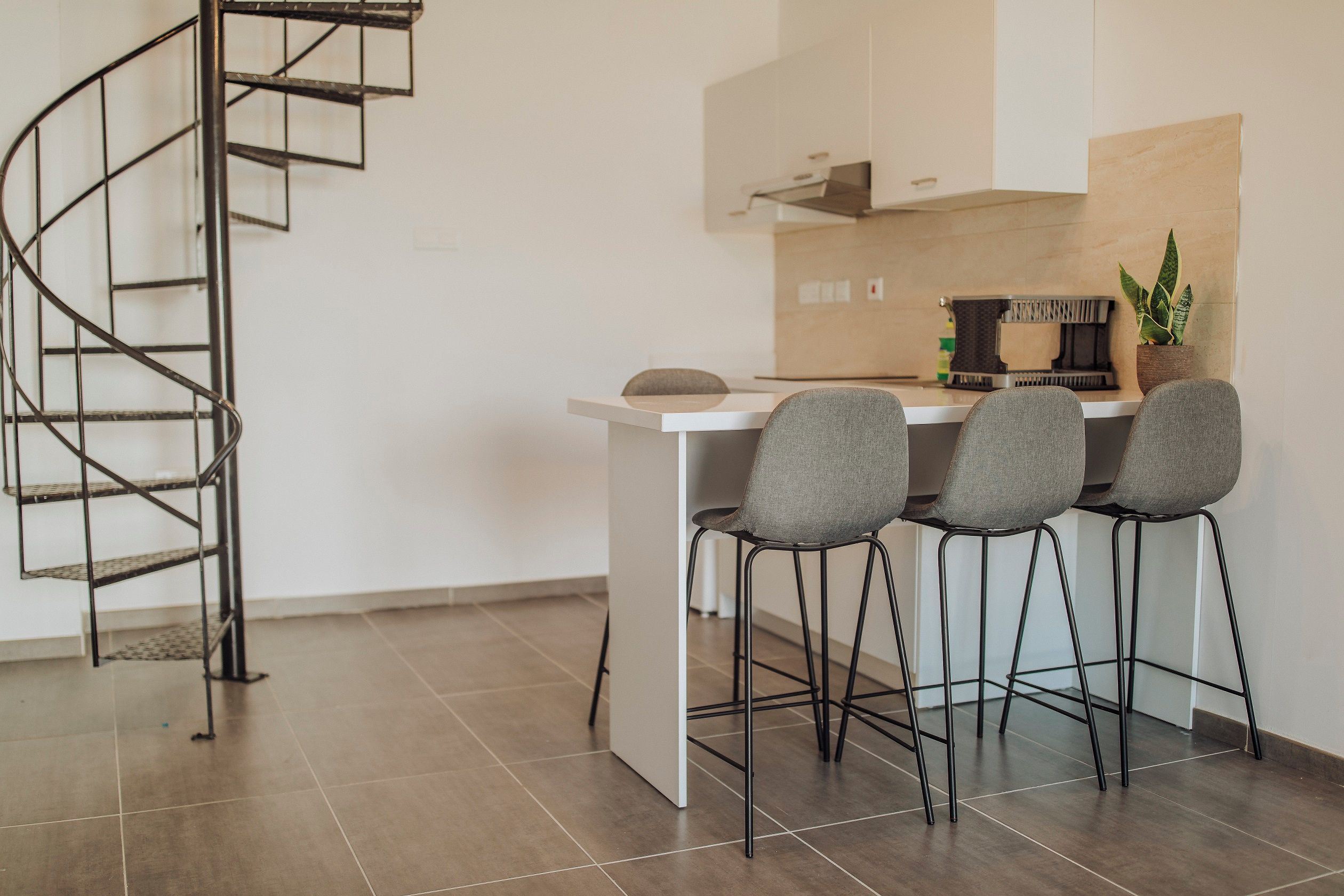 Eine moderne Küche mit weißer Arbeitsplatte, vier grauen Stühlen, einer schwarzen Metall-Wendeltreppe und einem Topf mit Pflanze auf der Arbeitsplatte.