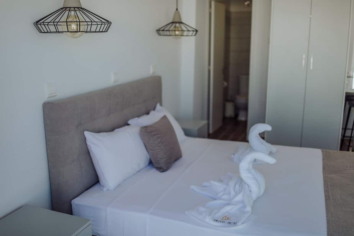 Cama bem arrumada com roupa de cama branca, cabeceira cinza e toalha em formato de cisne sobre a cama. Duas luminárias pendentes estão acima da cama, e um guarda-roupa é visível ao fundo.